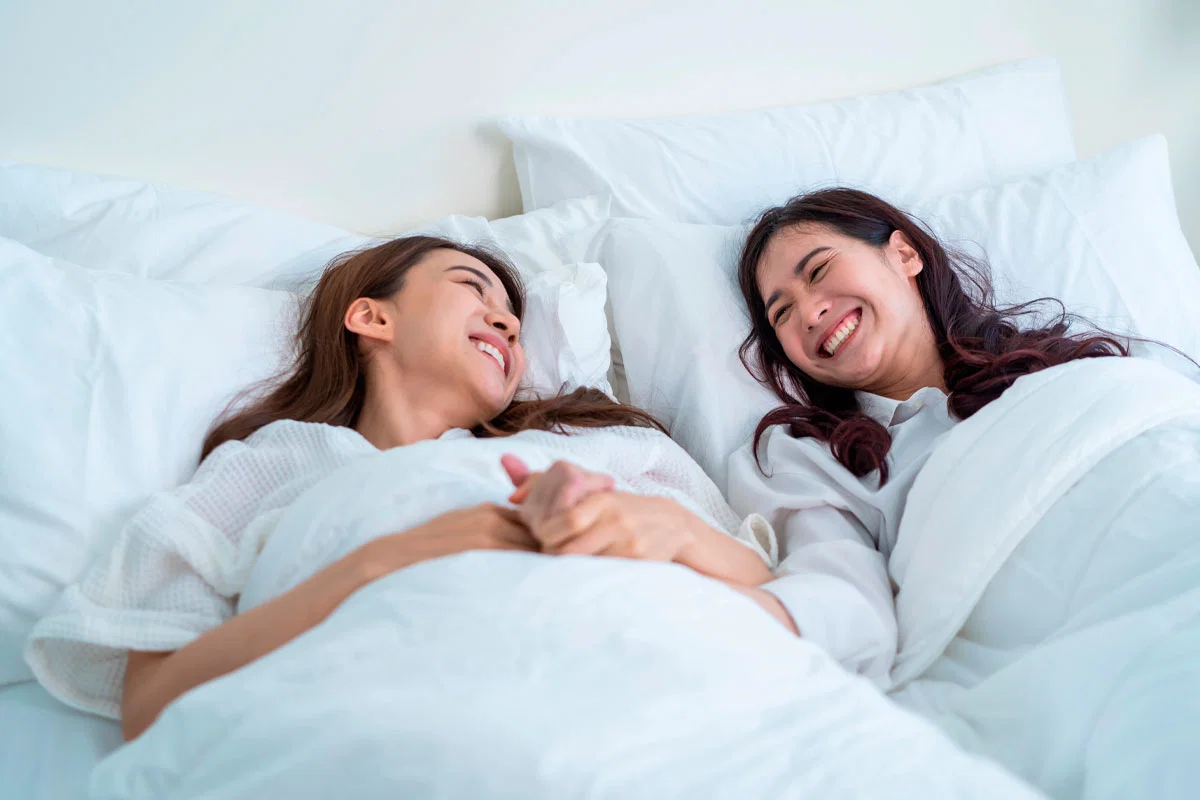 Dos mujeres se ríen tumbadas en la cama, tapadas con un edredón blanco y tomadas de la mano