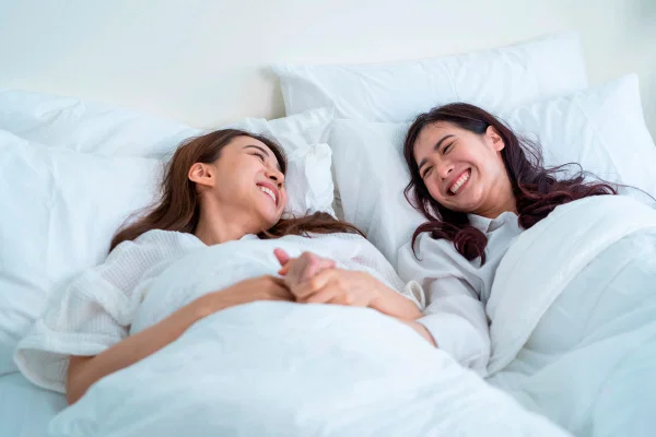 Dos mujeres se ríen tumbadas en la cama, tapadas con un edredón blanco y tomadas de la mano